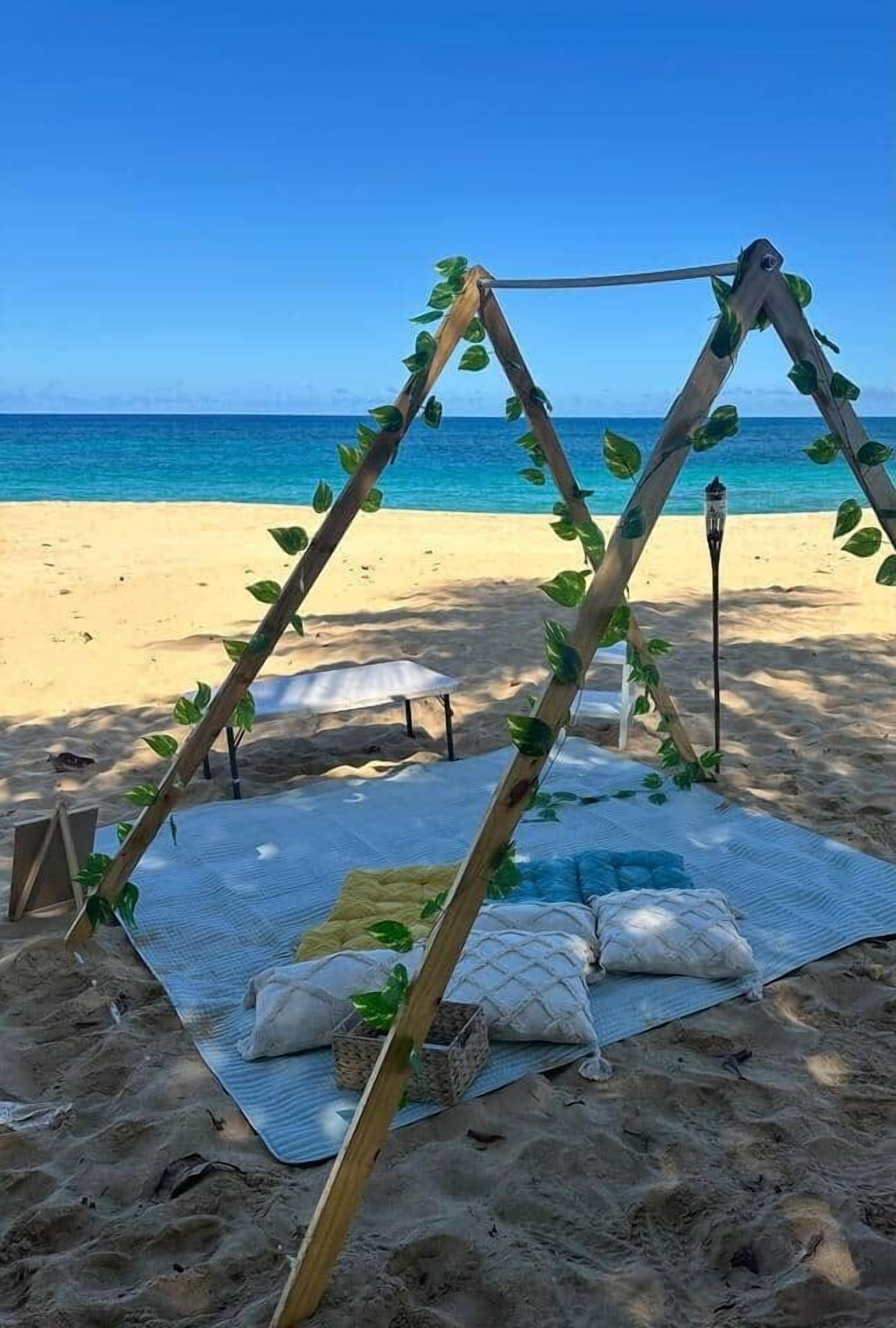 A beach setting with a wooden swing structure adorned with green foliage, a white mat with pillows, and a small table.