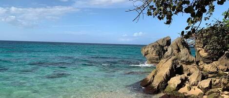 Ein Strand mit klarem türkisblauem Wasser, sandigem Ufer und Felsen.