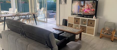 A modern living room with a grey sofa, a wooden coffee table, a TV on a white cabinet, and a sliding glass door leading to an outdoor area with chairs and a table.