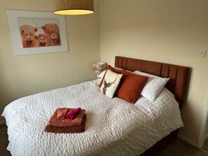 Una cama impecablemente hecha con una colcha blanca con textura, un cabecero marrón y almohadas decorativas, incluyendo una con un diseño de ciervo. Encima de la cama, un cuadro enmarcado de vacas, y una lámpara visible en la mesita de noche.