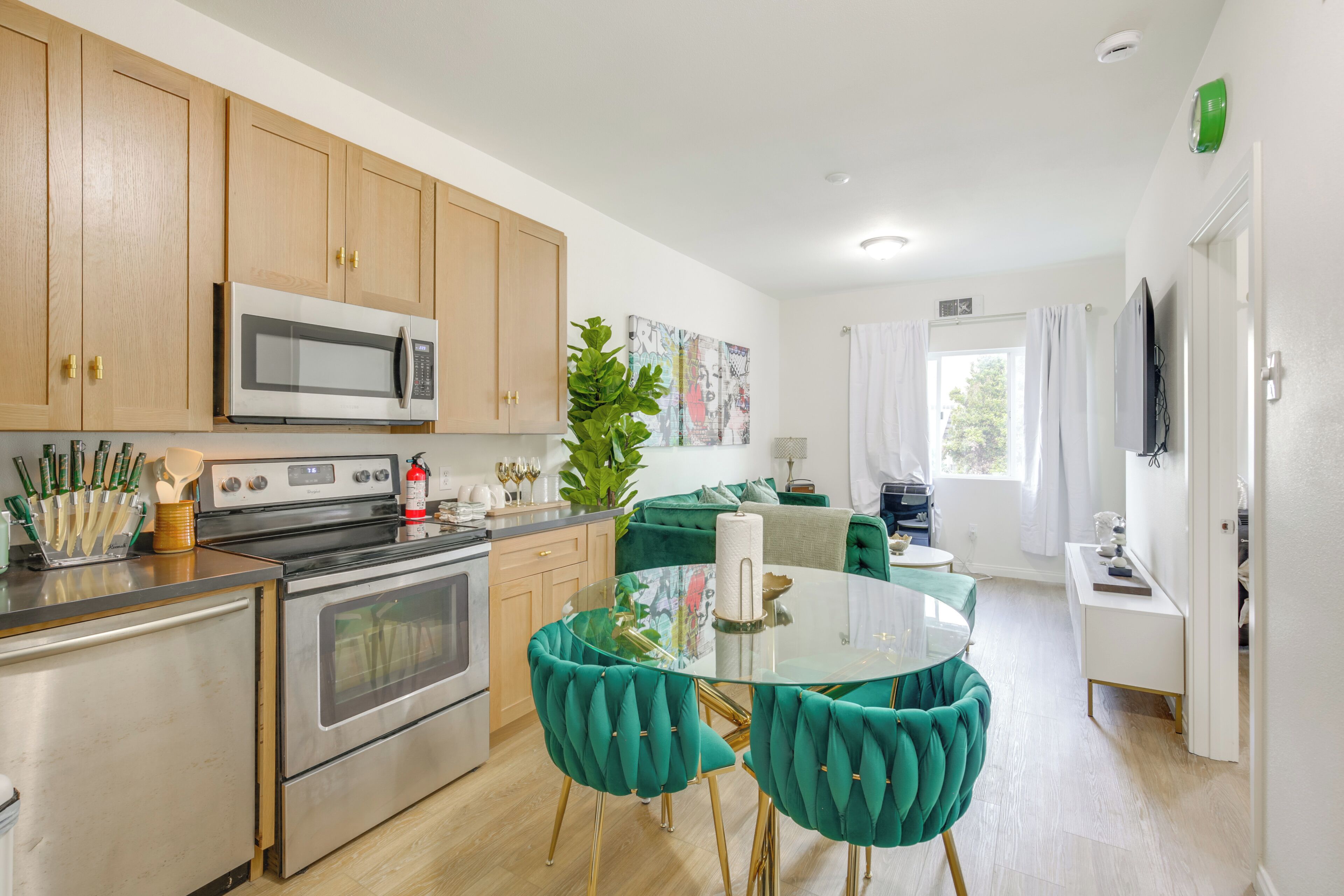 A modern kitchen with stainless steel appliances, a dining area with teal chairs, and a living space with a sofa and TV.