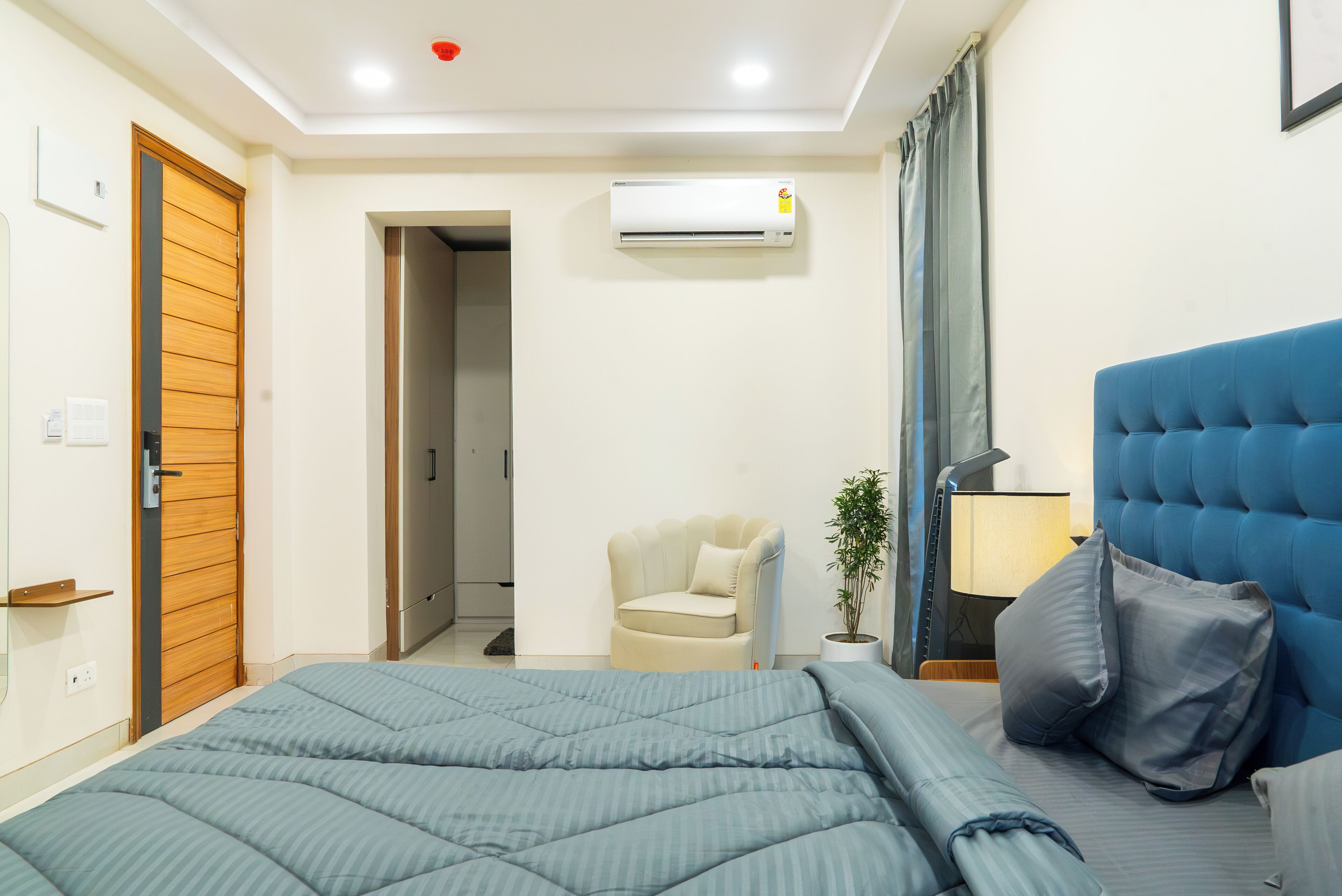 Quarto com cama, cabeceira azul, mesa de cabeceira, abajur, cadeira e ar condicionado.