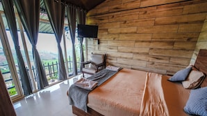 A bedroom with a wooden headboard, a bed with a beige bedspread, a chair, and a TV mounted on the wall. - Silent Valley Farmstead by VOYE HOMES (Ooty)