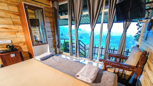 A wooden cabin room with a bed, a chair, a small table, and a window overlooking a scenic landscape. - Silent Valley Farmstead by VOYE HOMES (Ooty)