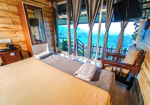 A wooden cabin room with a bed, a chair, a small table, and a window overlooking a scenic landscape. - Silent Valley Farmstead by VOYE HOMES (Ooty)
