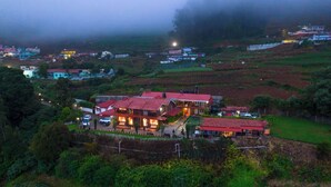 A large, multi-story building with a red roof, surrounded by greenery and fields. - Silent Valley Farmstead by VOYE HOMES (Ooty)