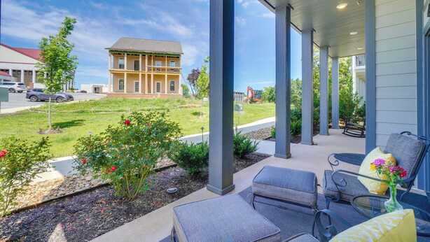A covered outdoor seating area with chairs and a table, overlooking a grassy lawn and a building in the background.