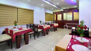 A restaurant with multiple tables, each set with white plates, red tablecloths, and chairs. - Hotel Indraprastha Kayamkulam (Kayamkulam)