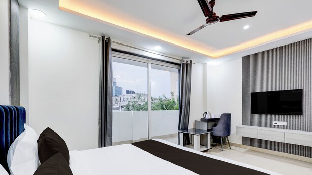 A modern hotel room with a large bed, a desk, a chair, a TV, and a balcony with a city view. - The Byke Business Hotel - Sector 29 (Gurugram)