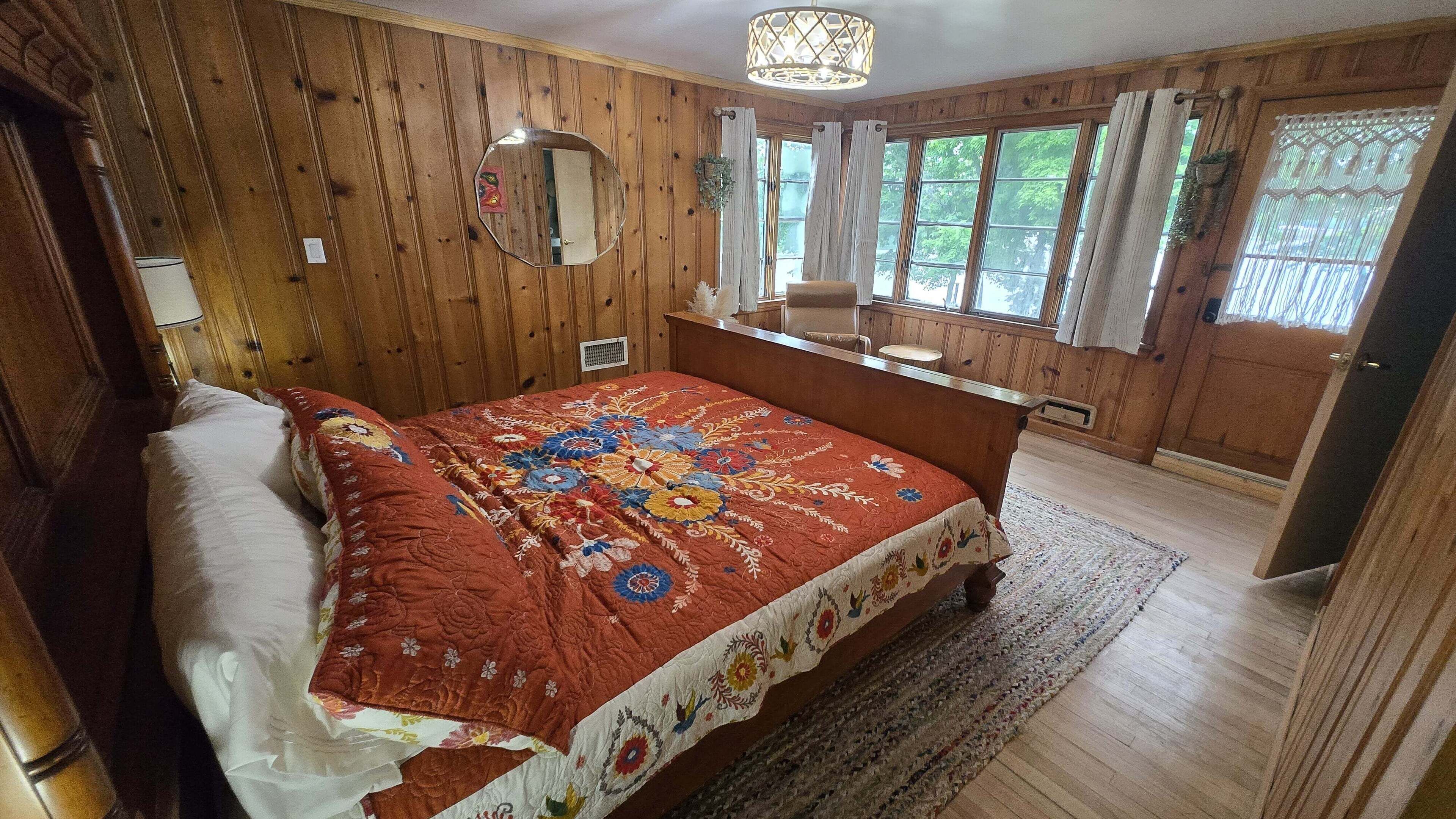 Une chambre avec un mur lambrissé en bois, un lit recouvert d’une courtepointe à motifs floraux, une table de chevet, un miroir et une fenêtre avec des rideaux.