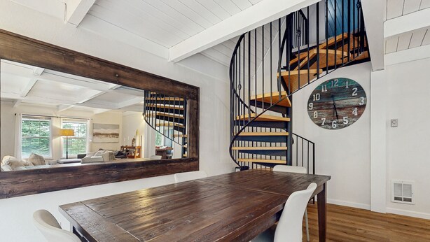 A modern dining area with a wooden table, white chairs, a large mirror, a spiral staircase, and a clock on the wall.