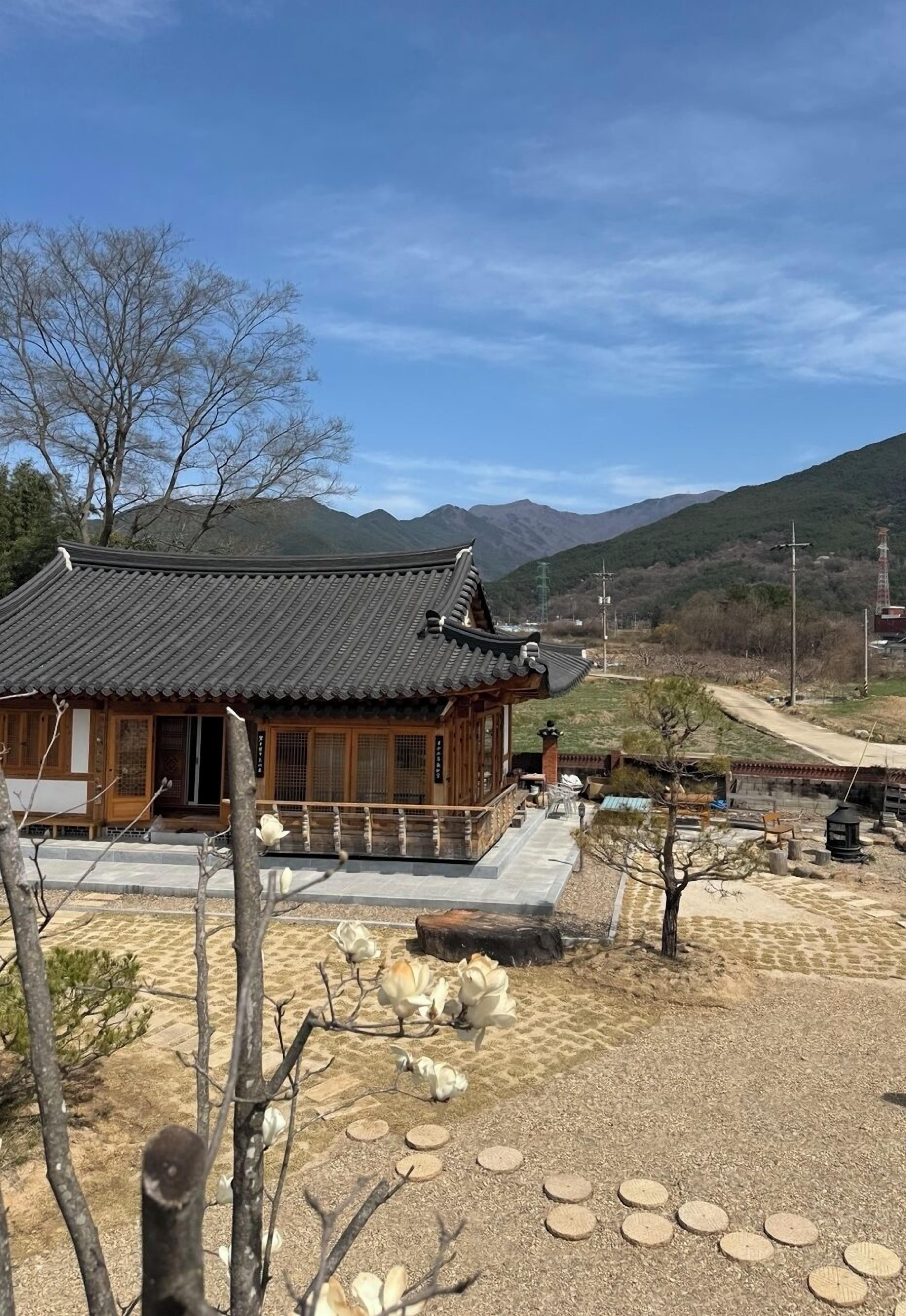 A traditional wooden building with a tiled roof, surrounded by a gravel courtyard with stepping stones and a few trees.