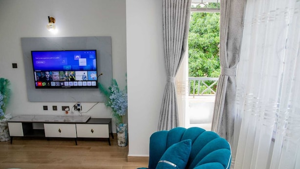 A living room with a flat-screen TV mounted on the wall, a teal armchair, a white console table, and a view of greenery through the window. - AZANIAHOMES (Nairobi)