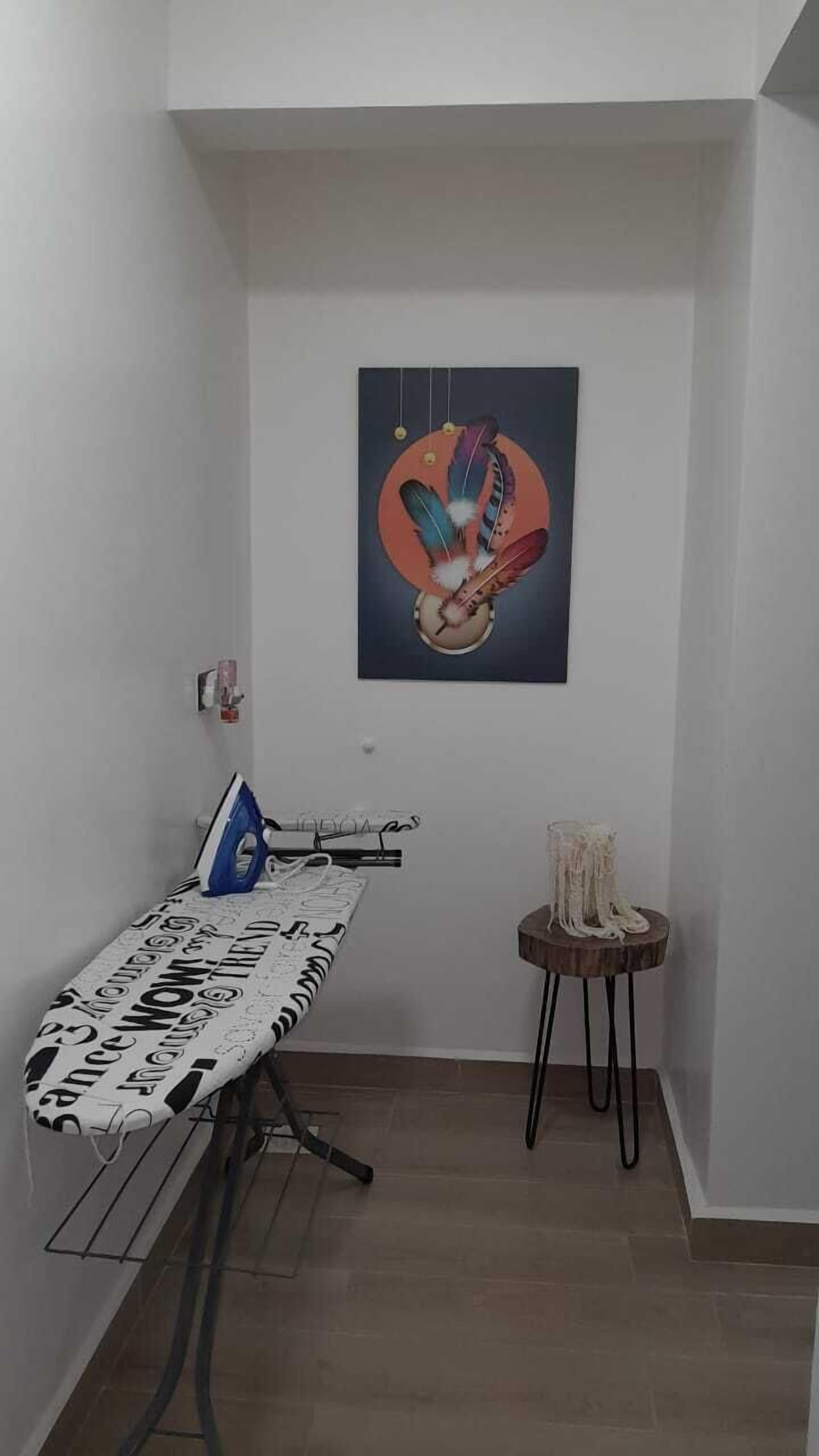 A corner room with a bed, an ironing board, a small table with a decorative item, and a wall-mounted painting.