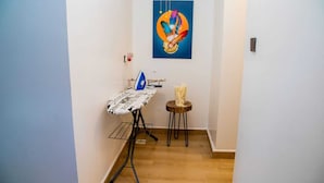 A narrow hallway with a wooden floor, an ironing board with an iron, a small round table with a decorative item, and a colorful painting on the wall. - AZANIAHOMES (Nairobi)