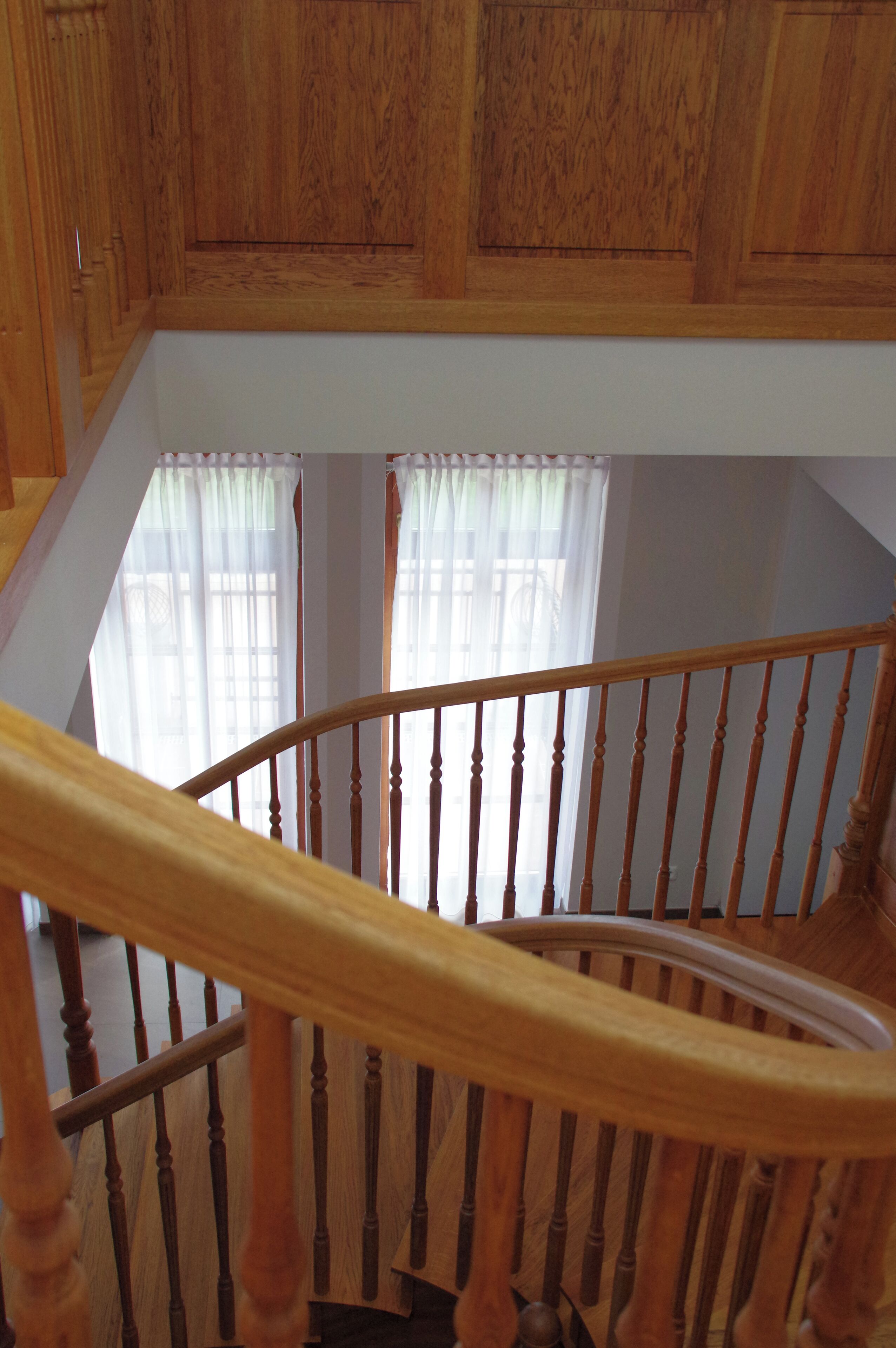 Un escalier en bois, doté d’une rampe, mène à un palier orné de lambris en bois et offrant une vue sur une fenêtre drapée de rideaux légers.