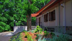 A building with a covered porch, a stone wall, and a garden with various plants and a tree. - Dwór Uradz (gmina Barwice)