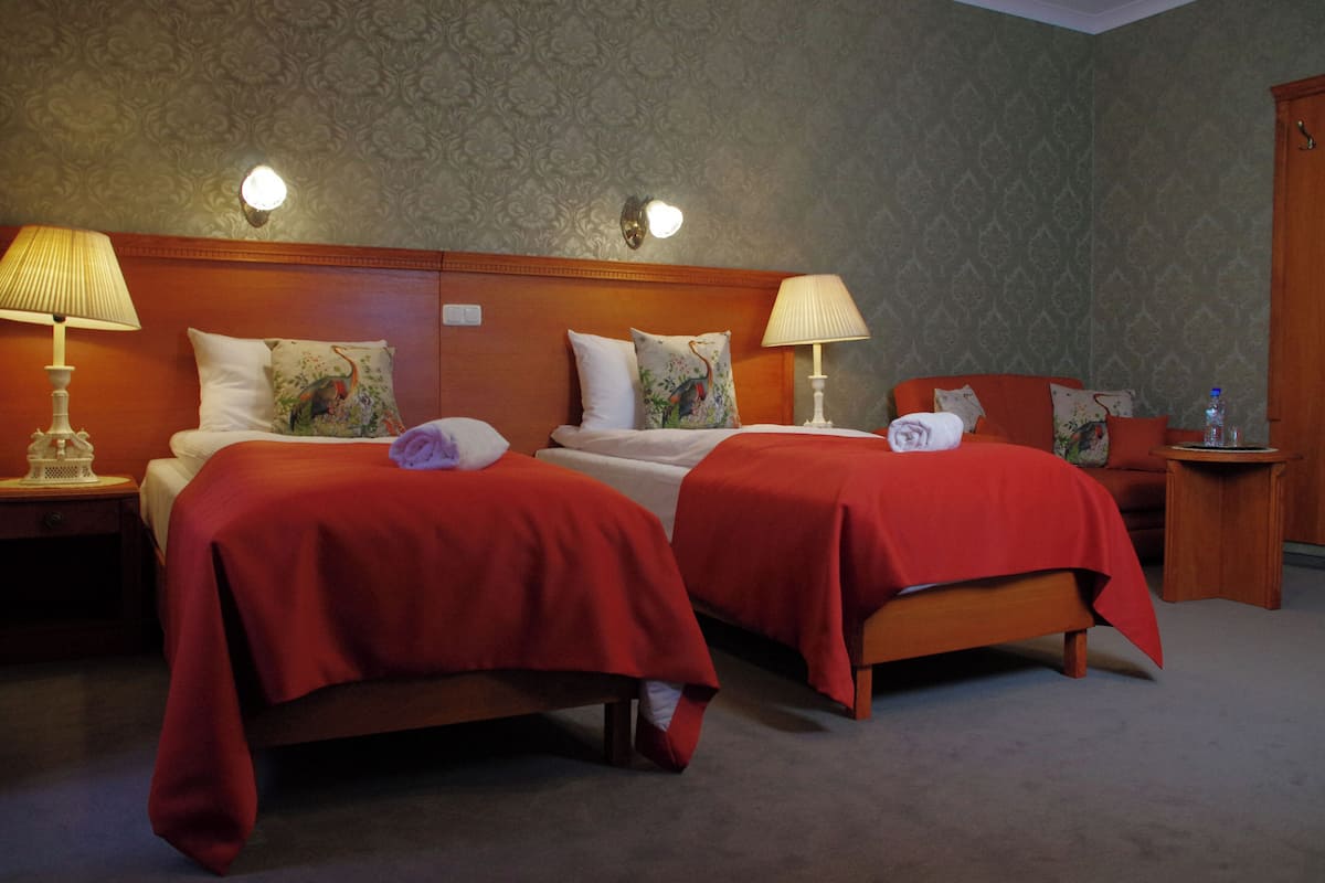 Habitación de hotel con dos camas, cada una con cubrecama rojo y almohadas blancas, cabecero de madera y dos lámparas fijadas a la pared.