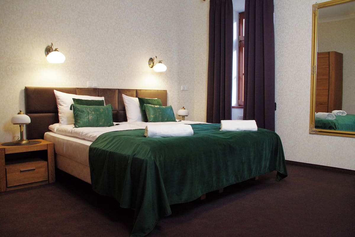 Una cama doble con ropa de cama verde, dos mesitas de noche con lámparas, un espejo grande y una ventana con cortinas.