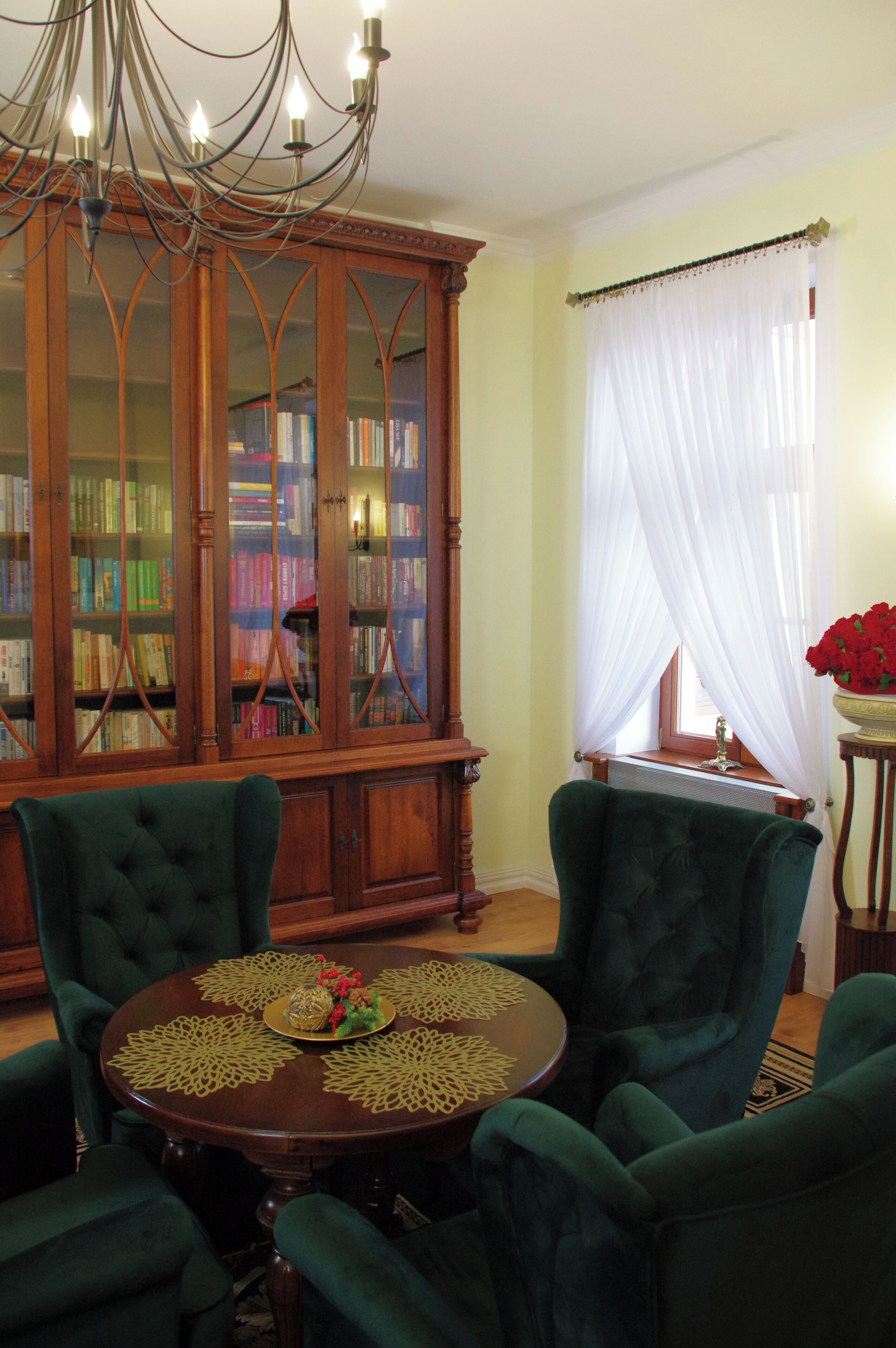 Um quarto com uma estante de madeira, uma mesa redonda com cadeiras verdes e um lustre.