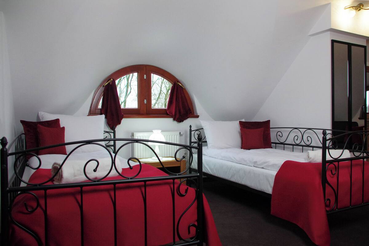 Habitación con dos camas, cada una con colcha roja y ropa de cama blanca. Se ve una ventana con marco de madera y cortina roja.