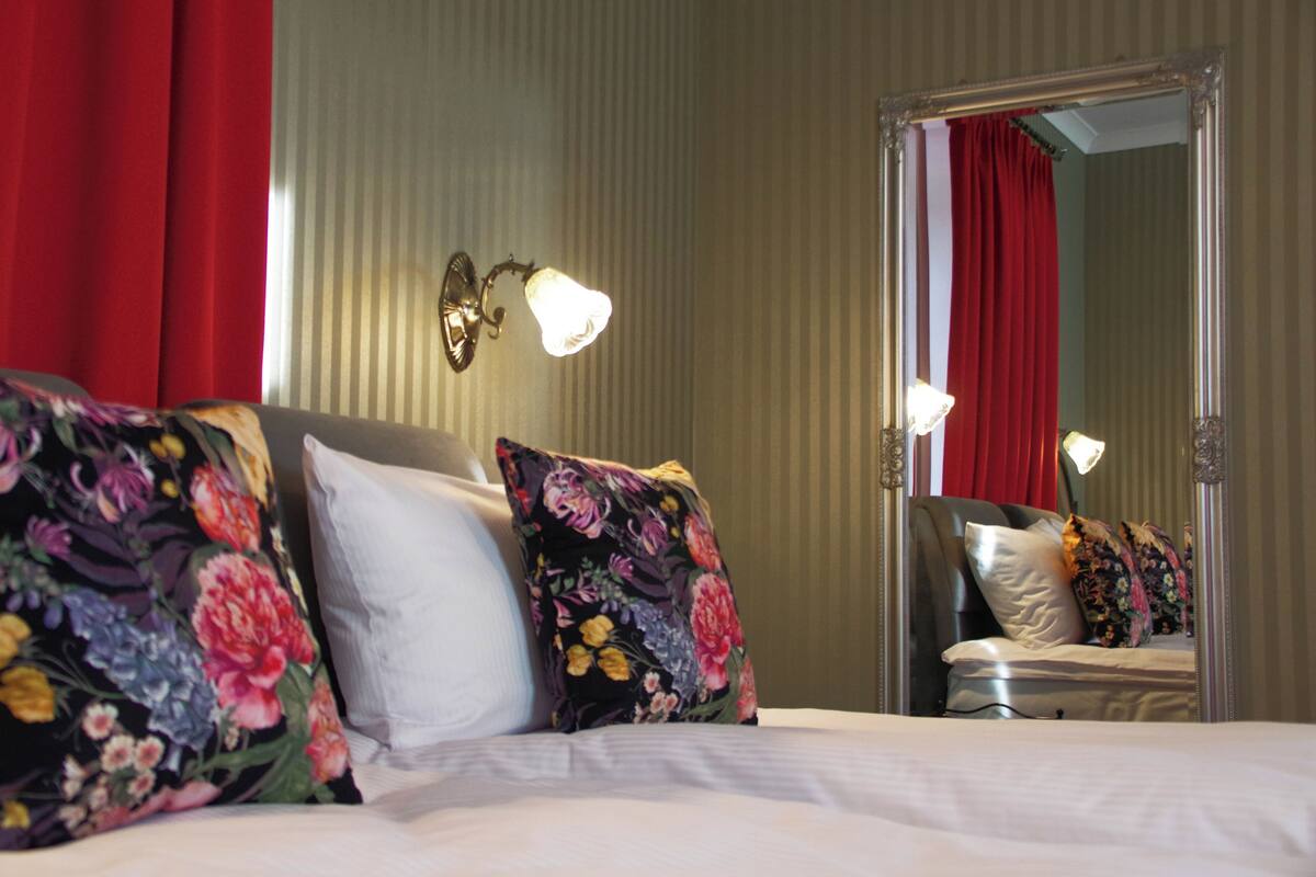 Una habitación de hotel con una cama, almohadillas estampadas con flores, un espejo y una cortina roja.