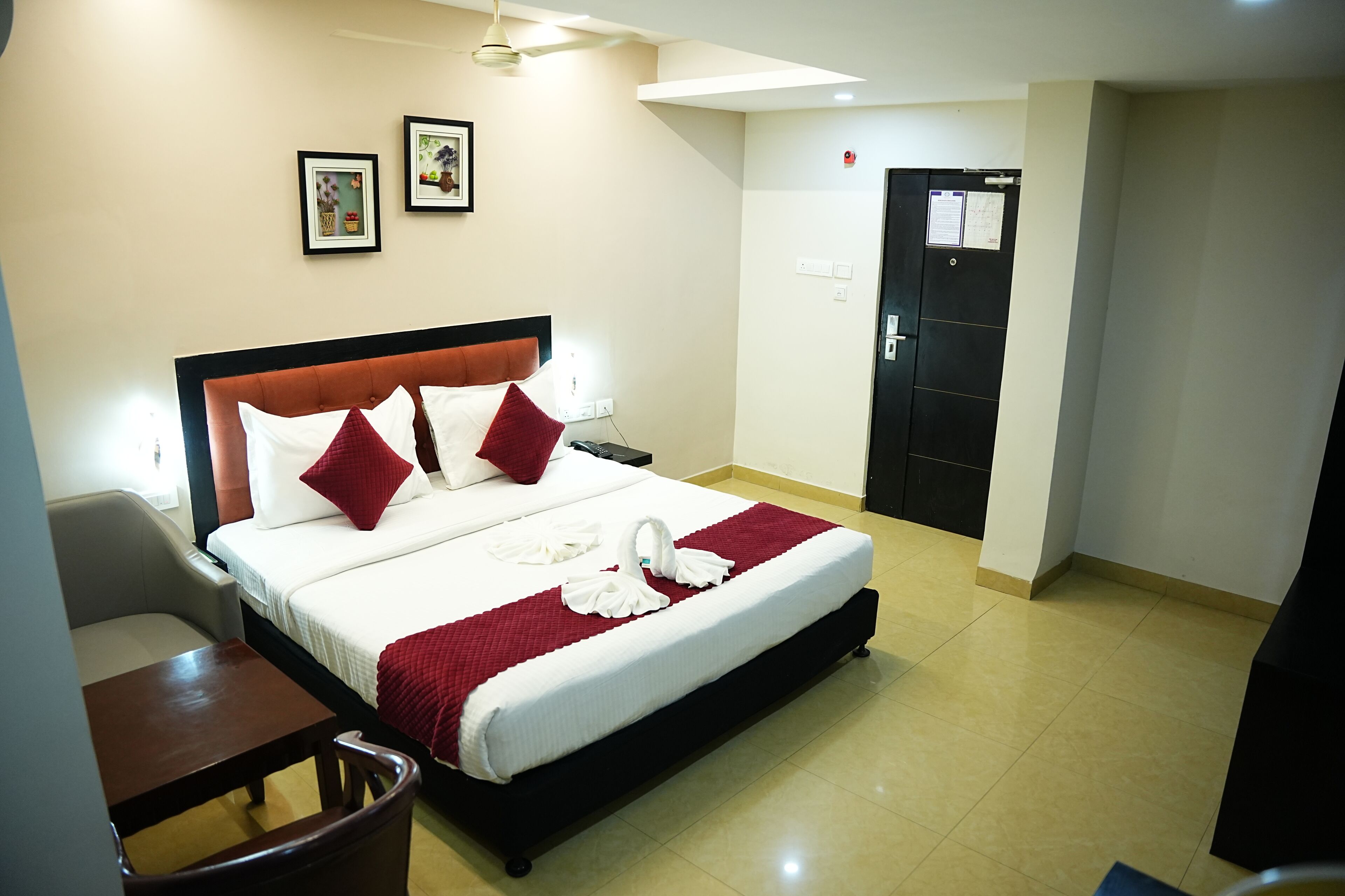 A hotel room with a bed, a desk, and a chair. There is a door and a wall with framed pictures.