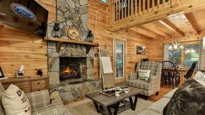 A cabin interior with a stone fireplace, wooden walls, a dining area, and a cozy seating arrangement.