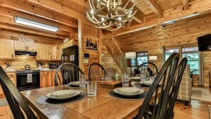 A wooden cabin interior with a dining table set for four, a kitchen with wooden cabinets, a chandelier, and a staircase.