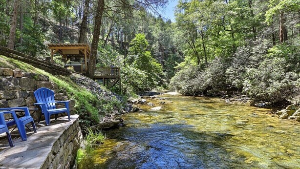 A clear stream with a wooden deck and blue chairs, surrounded by trees.