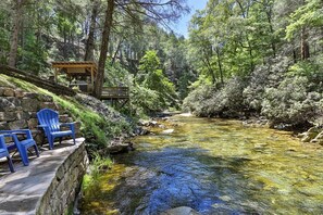 Un ruisseau aux eaux claires, avec une terrasse en bois et des chaises bleues, le tout entouré d’arbres.
