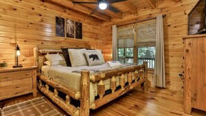 A wooden cabin bedroom with a bed, a nightstand, a ceiling fan, and a window with curtains.