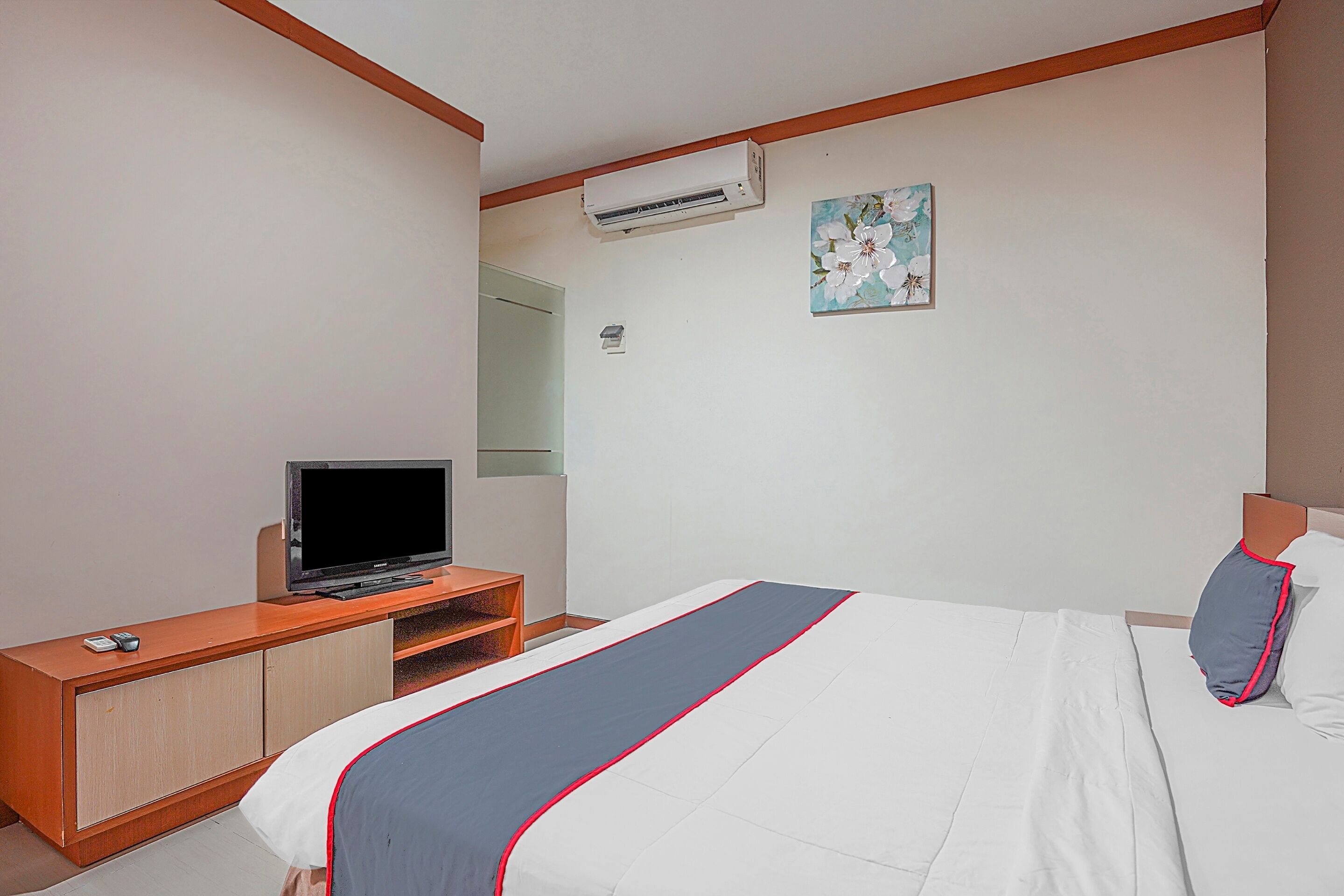 Una habitación de hotel con una cama, un televisor sobre un mueble de madera, un aire acondicionado y un cuadro en la pared.
