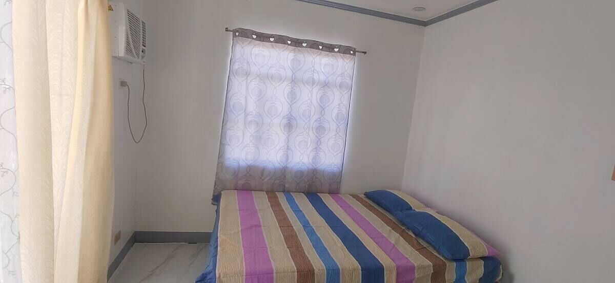A small, single-bed room with a striped bedspread, a window with a curtain, and a wall-mounted air conditioning unit.