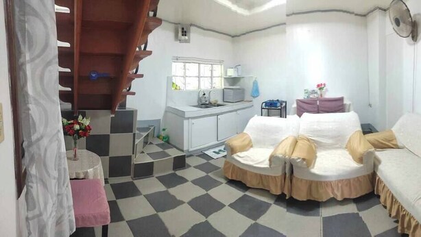 Una habitación con suelo a cuadros, un sofá, una silla, una mesita con un jarrón y una zona de cocina al fondo.