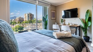 A hotel room with a large bed, a desk with a chair, a flat-screen TV, and a view of a cityscape through large windows. - M&C Departamento Irarrazaval 614 (Santiago)