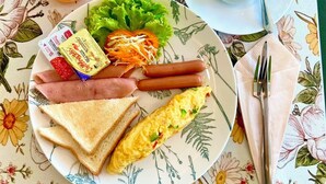 Um prato com torradas, presunto, salsichas, alface, cenoura ralada e um omelete, acompanhado de um copo de suco de laranja e uma xícara de café preto sobre uma toalha de mesa florida.