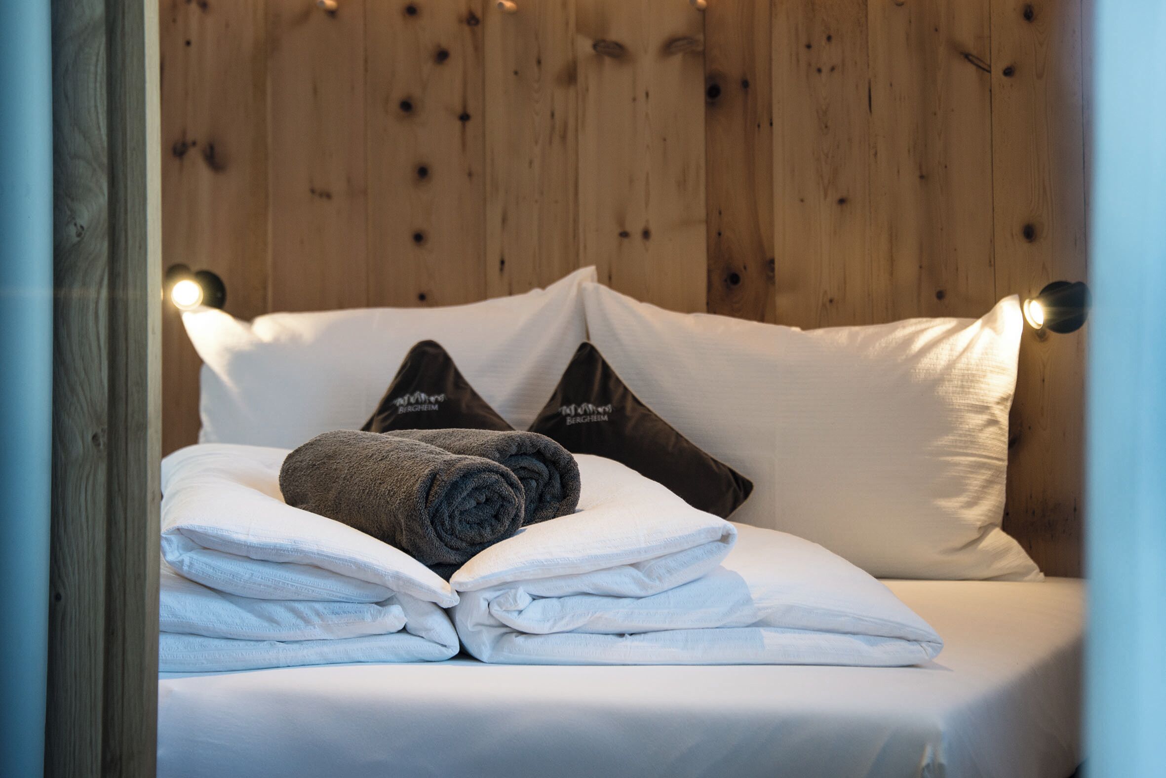 A neatly made bed with folded white towels and rolled towels, set against a wooden wall with a light fixture.
