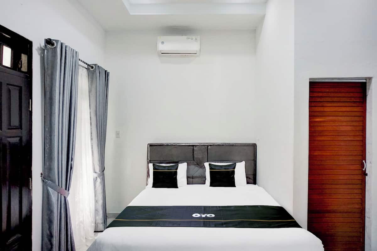 Uma cama de solteiro com cabeceira, janela com cortinas, ar condicionado instalado no teto e porta de madeira.