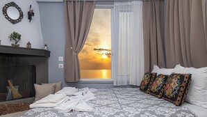 A bedroom with a large bed, a fireplace, and a view of a sunset through a window with curtains. - Barbara's Apartment Sea View (Stegna)