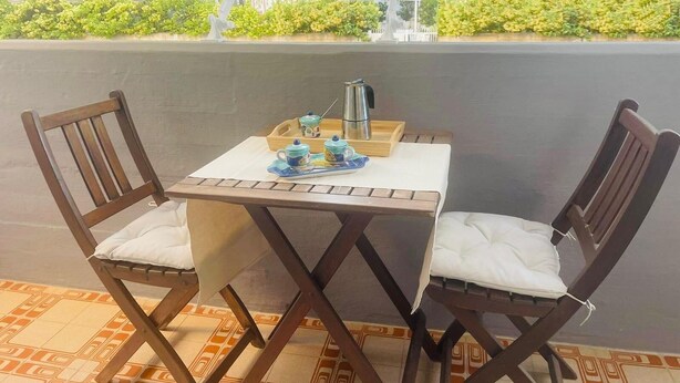 Une petite table d’extérieur avec deux chaises en bois, un plateau sur lequel se trouvent une théière, des tasses et des soucoupes, et une vue sur un jardin verdoyant par la fenêtre.