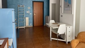 Une pièce avec un mur bleu, une porte en bois, un bureau blanc avec deux chaises, un réfrigérateur bleu et un meuble blanc.