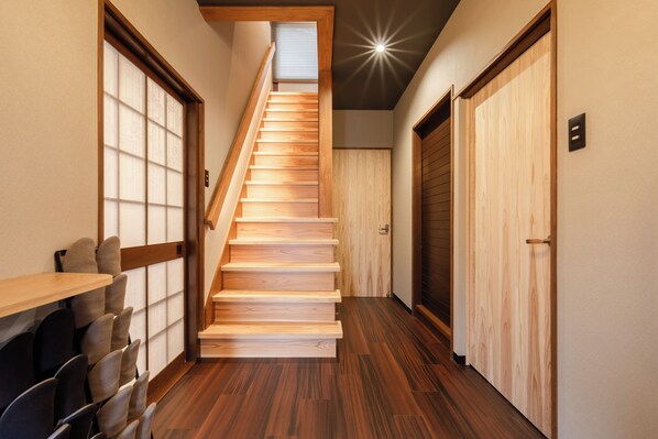 A wooden staircase in a hallway with a ceiling light, wooden doors, and a seating area with chairs.