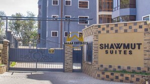 Une entrée fermée par un portail mène à un immeuble de grande hauteur, sur lequel on peut lire l’inscription « HAVAN COTTAGES SHAWMUT SUITES ».
