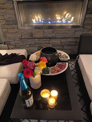 A cozy outdoor seating area with a stone wall backdrop, a fire feature, and a table set with food, flowers, and candles. - Rooftop Oasis over looking Shannon lake golf course (West Kelowna)