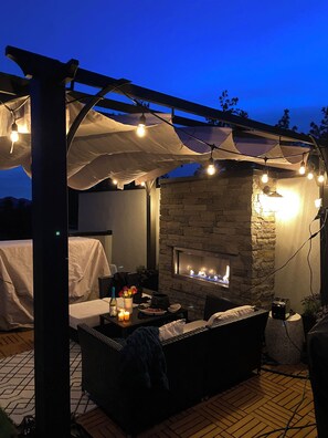 An outdoor patio area with a stone fireplace, string lights, and a covered seating area. - Rooftop Oasis over looking Shannon lake golf course (West Kelowna)