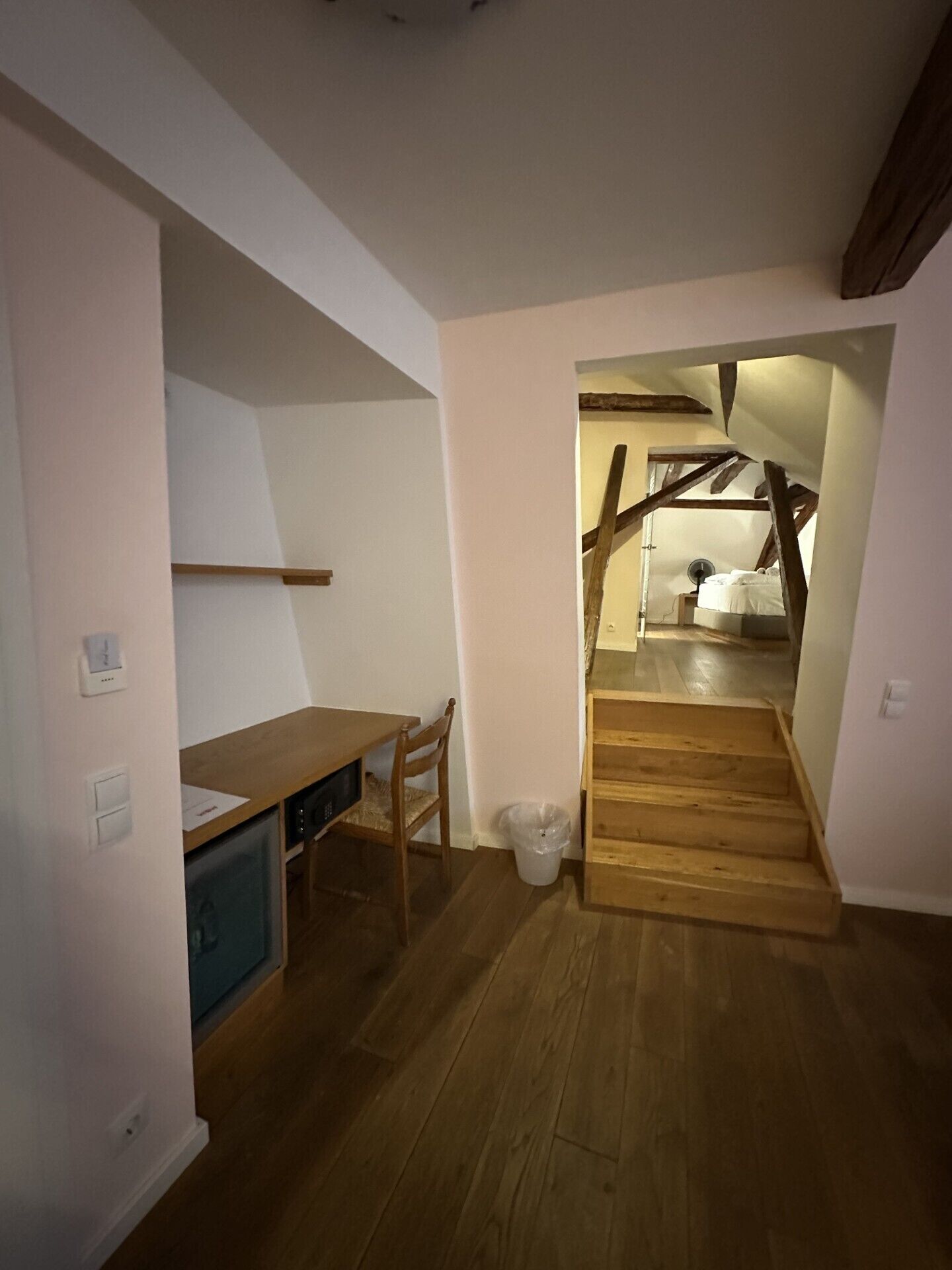 A compact room with a wooden desk, chair, and a small kitchenette area. A staircase leads to an upper level.