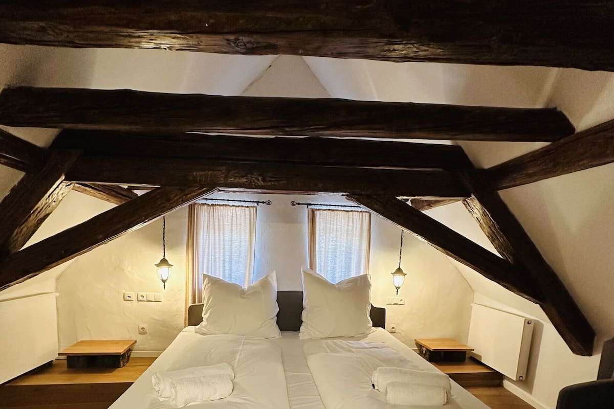 Ein Doppelbett in einem Loftsraum mit freiliegenden Holzbalken, zwei Nachttischen und einer hängenden Leuchte.