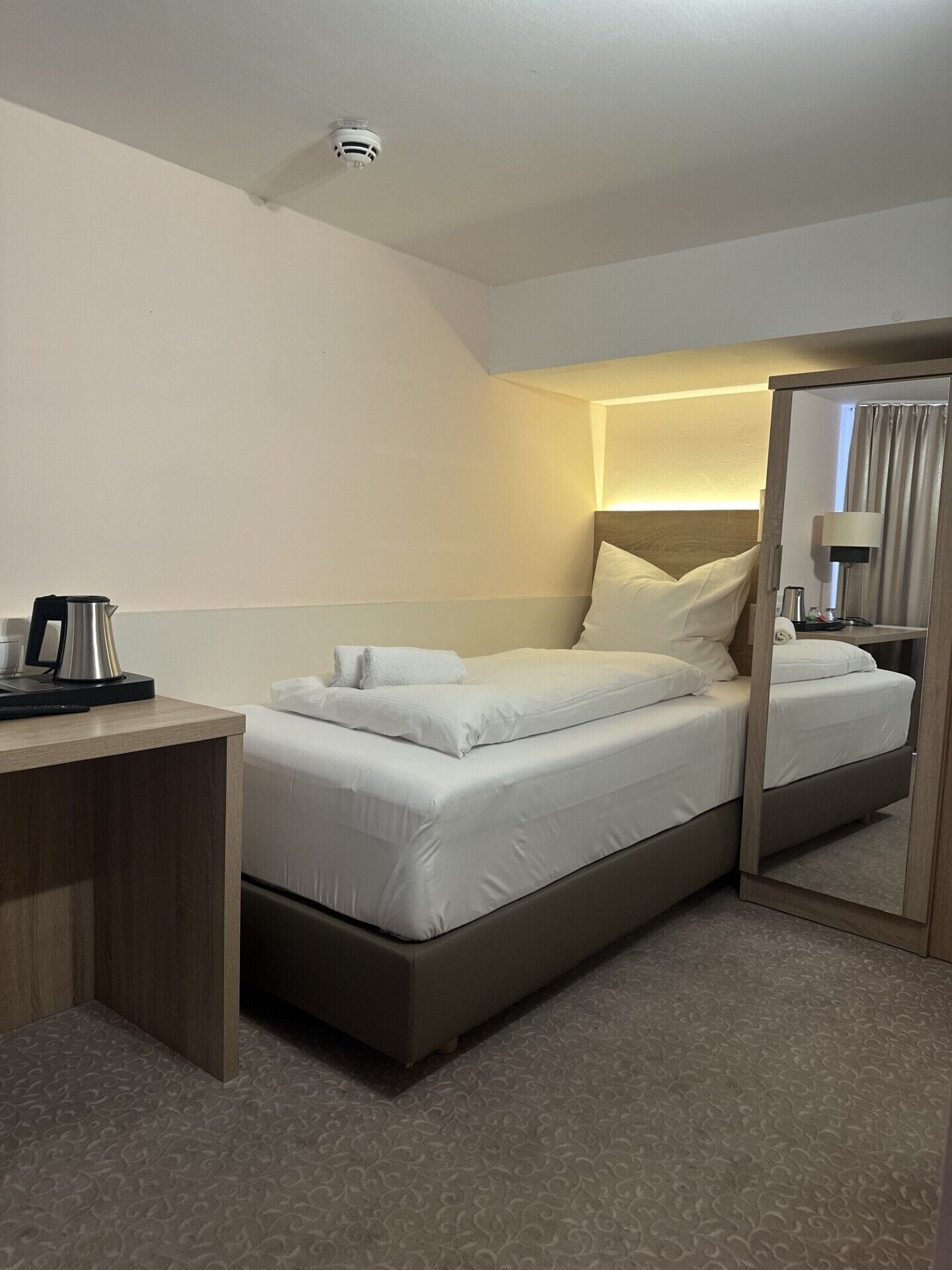 A hotel room with a bed, a desk with a coffee maker, a mirror, and a nightstand.