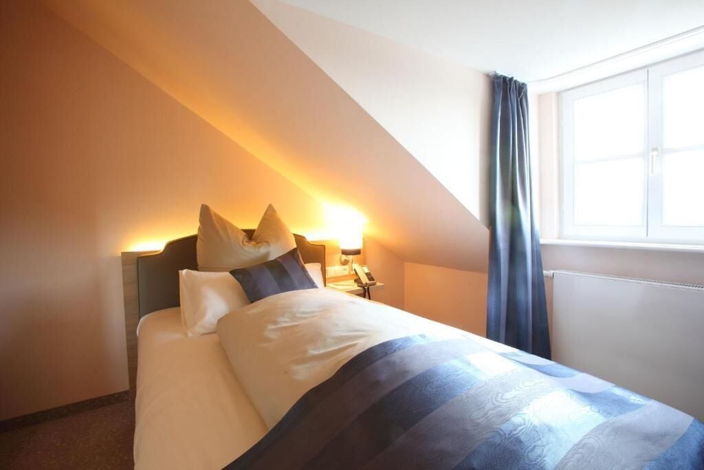 Una habitación de hotel con cama, mesita de noche, lámpara y ventana con cortinas.